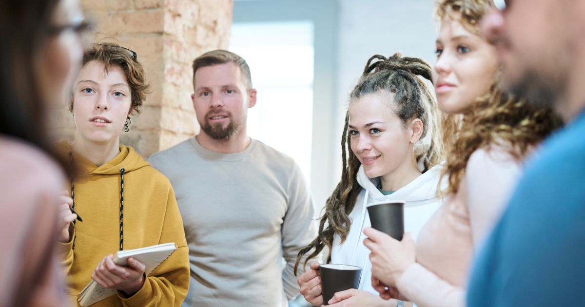 Four coworkers in hoodies and sweaters stand near a brick wall holding coffee cups and a notebook, listening to a colleague in the blurred foreground.