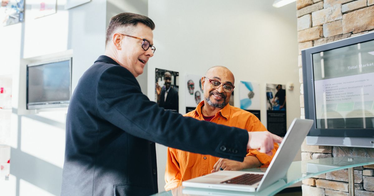 A man in glasses and a dark jacket smiles as he points at a laptop screen, showing it to a smiling colleague in an orange shirt at a glass desk.