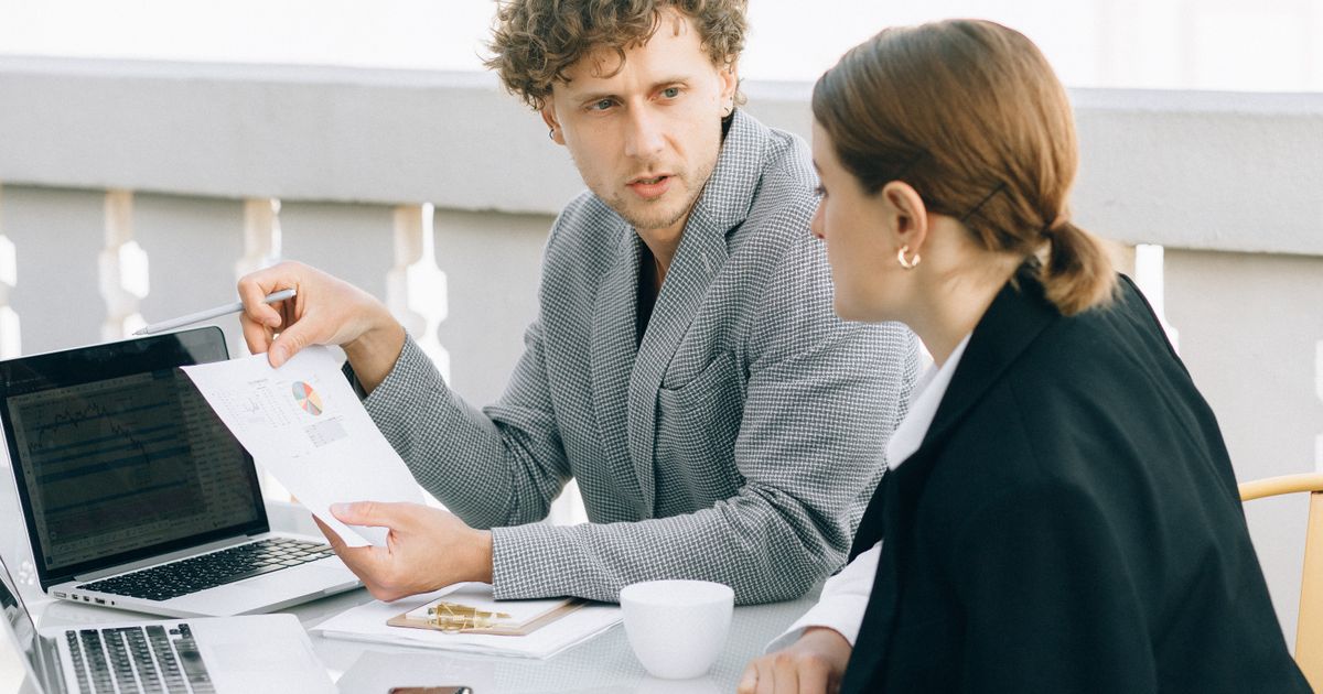 A man with curly hair in a checked jacket holds up a printed chart and explains it to a colleague with her hair tied back, at a table with laptops and a coffee cup.