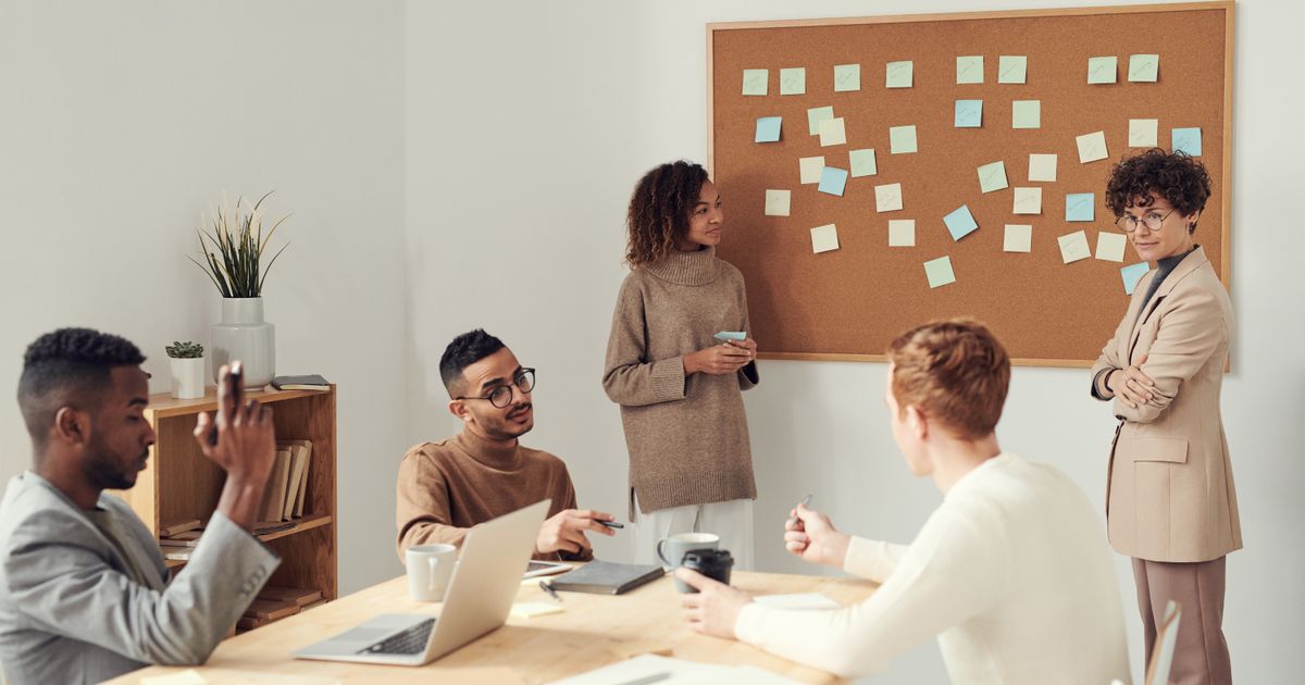 Two women stand beside a corkboard covered in pale sticky notes while three seated colleagues at a table with laptops and coffee cups talk with them.