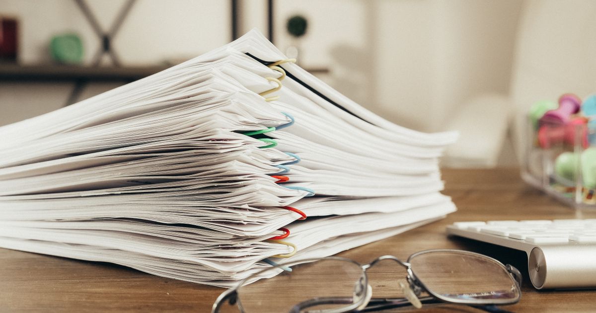 A tall stack of paper documents held together with colored binder clips sits on a wooden desk beside a pair of glasses and a keyboard.
