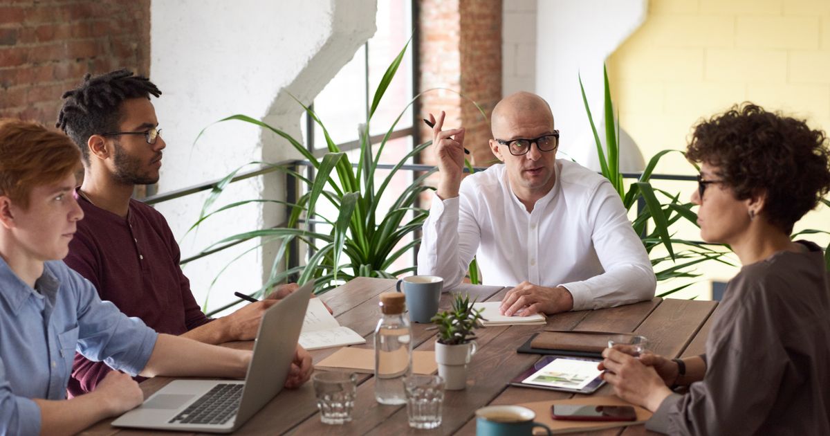 Four people sit around a wooden table with laptops and water glasses while a bald man in a white shirt talks and gestures, with plants behind him.