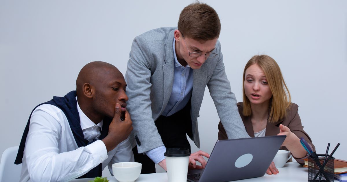Three colleagues lean over one laptop on a white desk, one with his hand over his mouth, studying the screen.