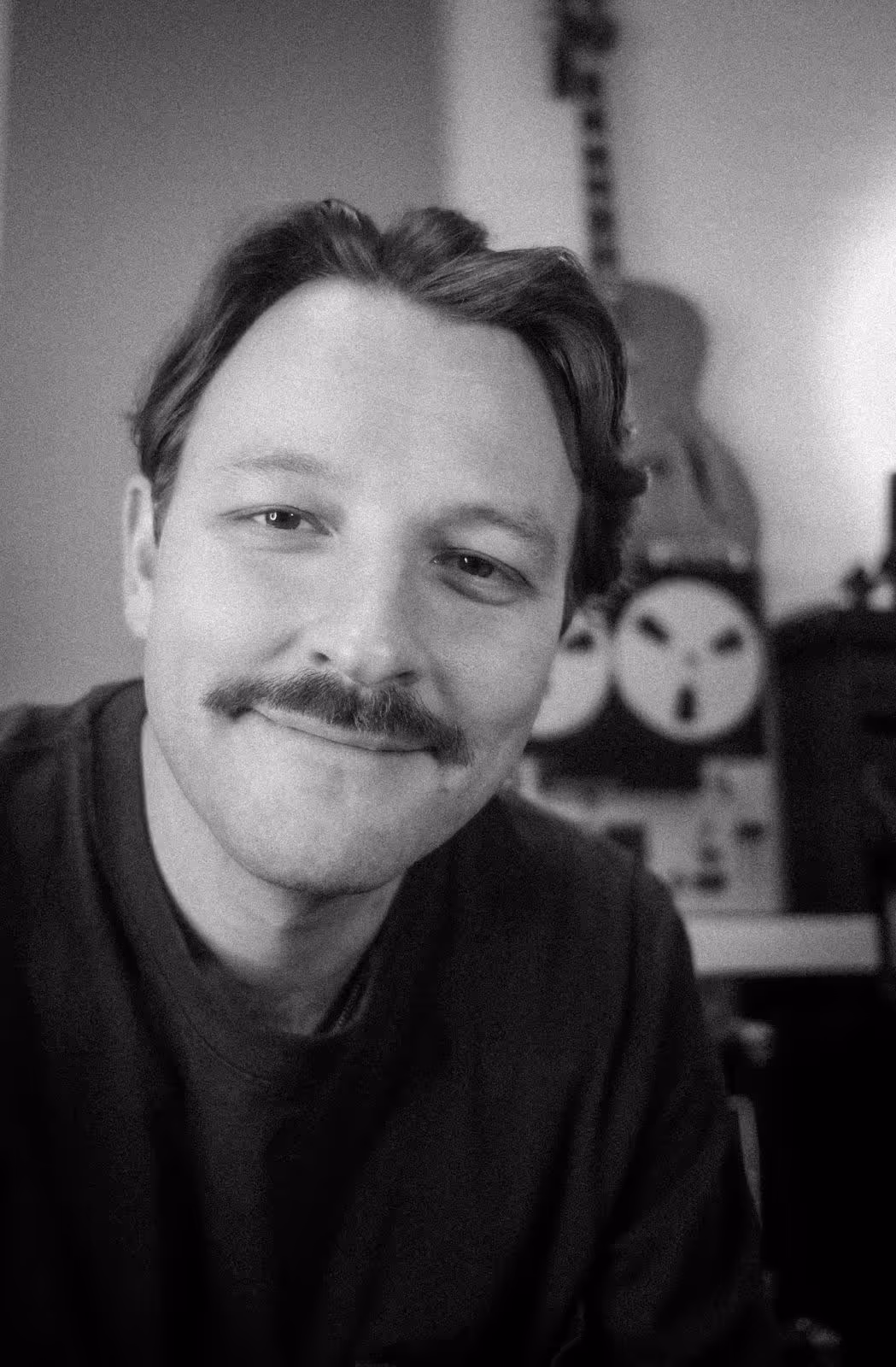 Black and white portrait of a man with a mustache smiling, with a guitar and round objects in the blurred background.