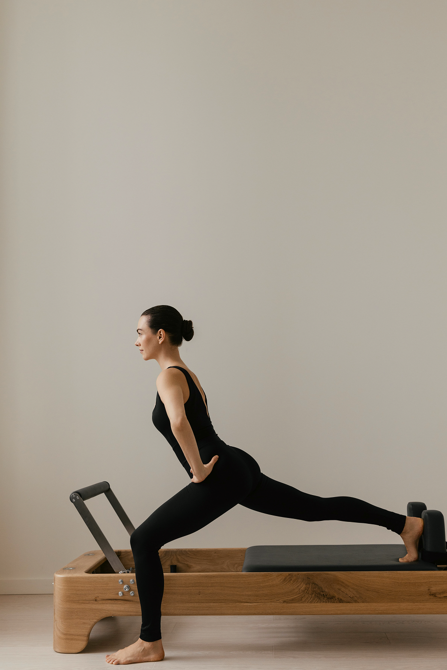 Woman in black workout attire doing a lunge stretch on a wooden Pilates reformer machine against a plain beige wall.