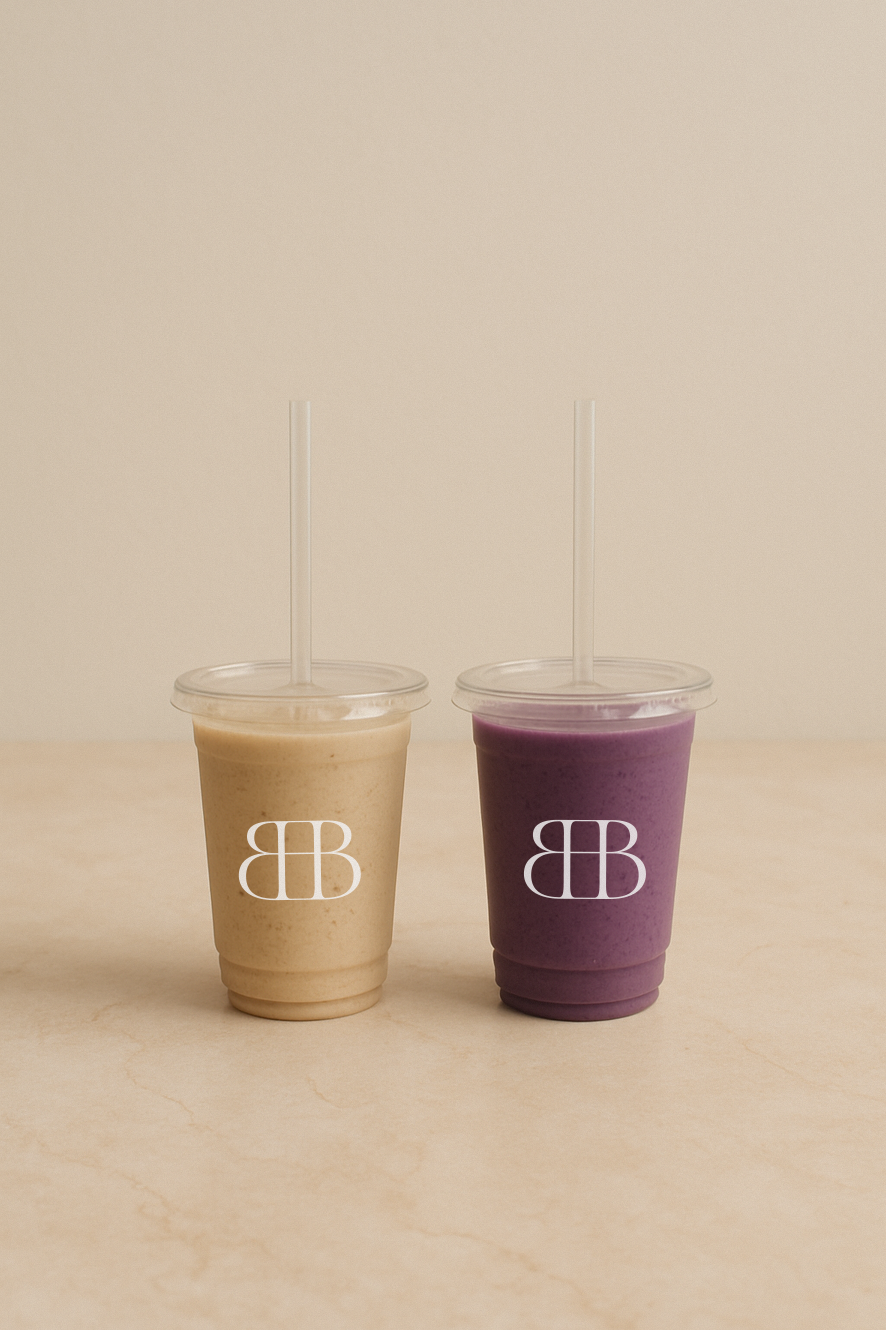 Two plastic cups with lids and straws containing beige and purple smoothies on a beige surface.