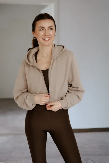 Smiling woman wearing a beige hoodie over a dark brown outfit standing indoors.