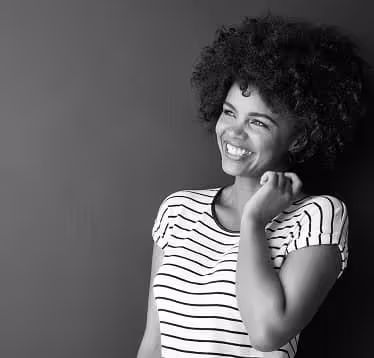 Smiling woman with curly hair wearing a striped shirt, looking to the side against a plain dark background.