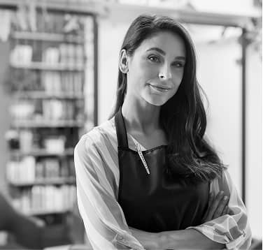 Confident woman with long dark hair wearing an apron, standing with folded arms in a room with bookshelves in the background.