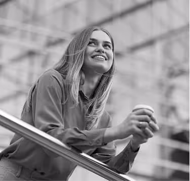 Smiling woman holding a coffee cup while leaning on a railing indoors.