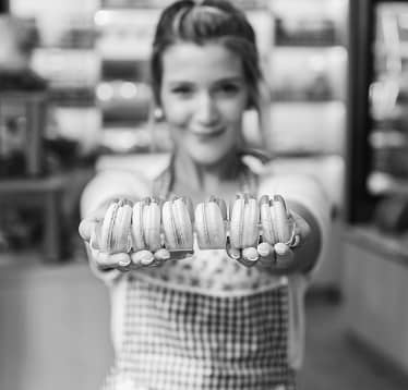 Woman in apron holding out a row of macarons towards the camera in a bakery.