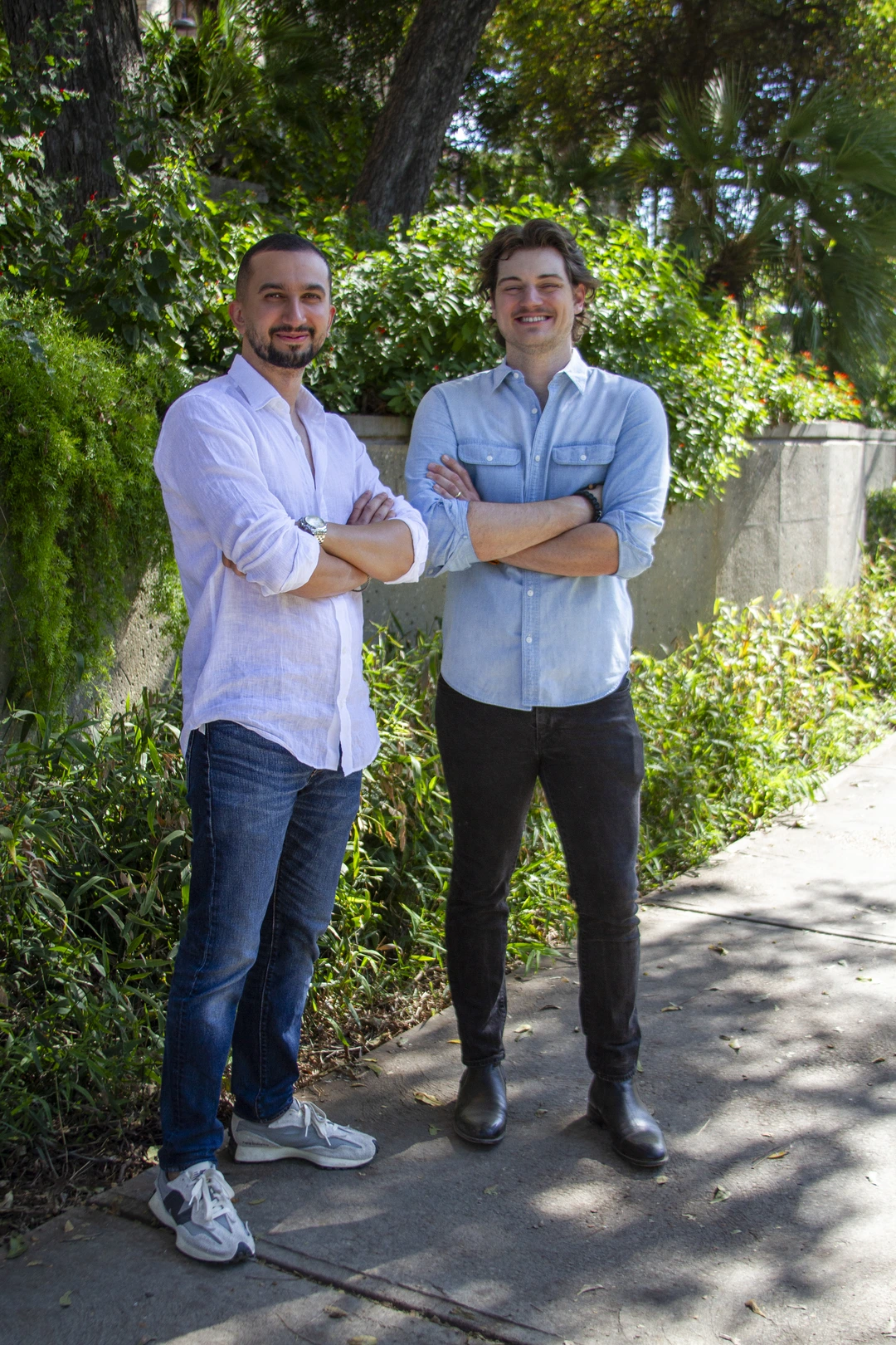 Two men standing outdoors with arms crossed, smiling in front of greenery and trees.