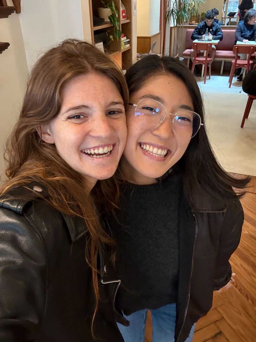 Two young women smiling closely together indoors, one with glasses and the other with long brown hair, both wearing dark jackets.