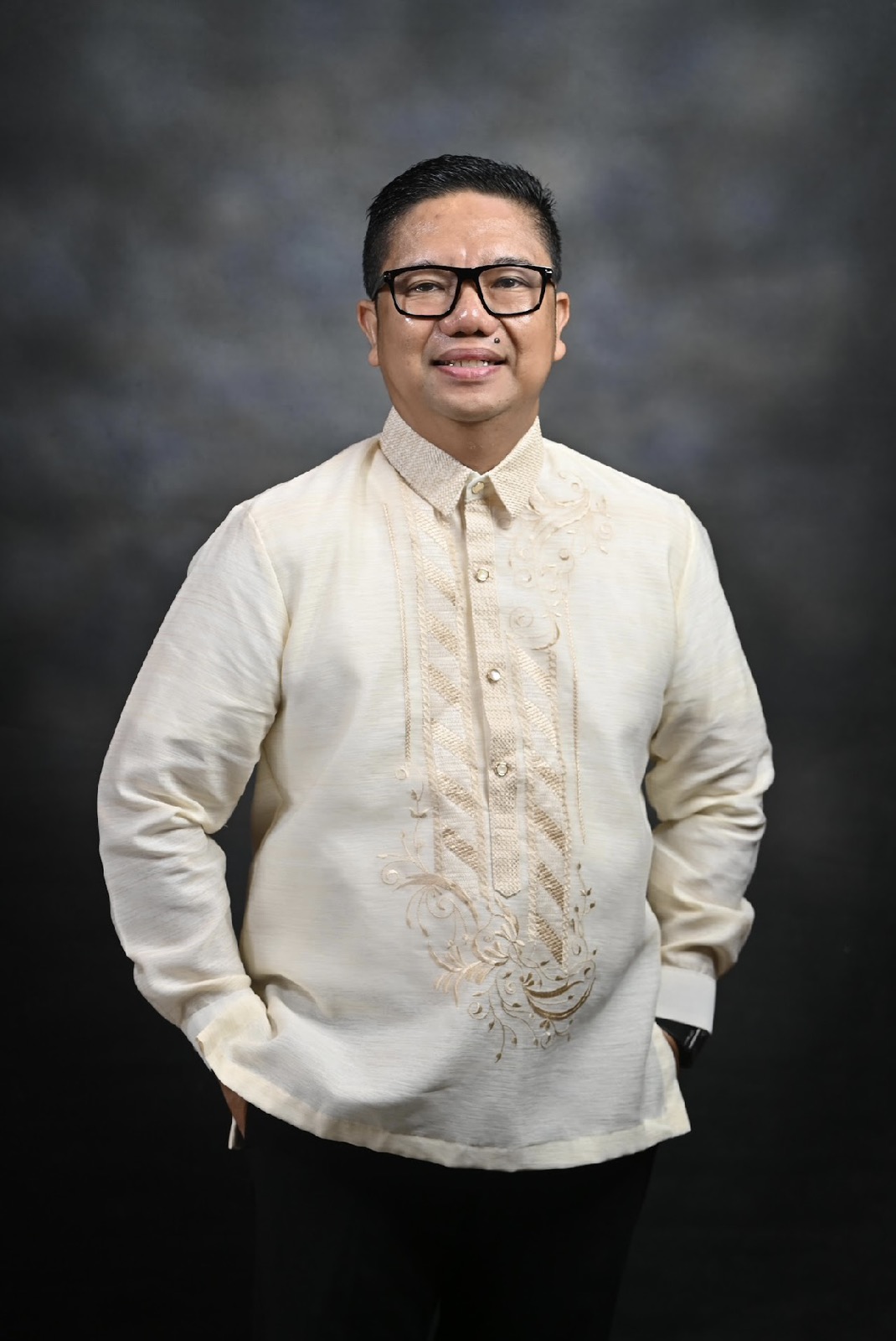 Man wearing traditional Filipino Barong Tagalog, smiling with hands in pockets against a dark gray backdrop.