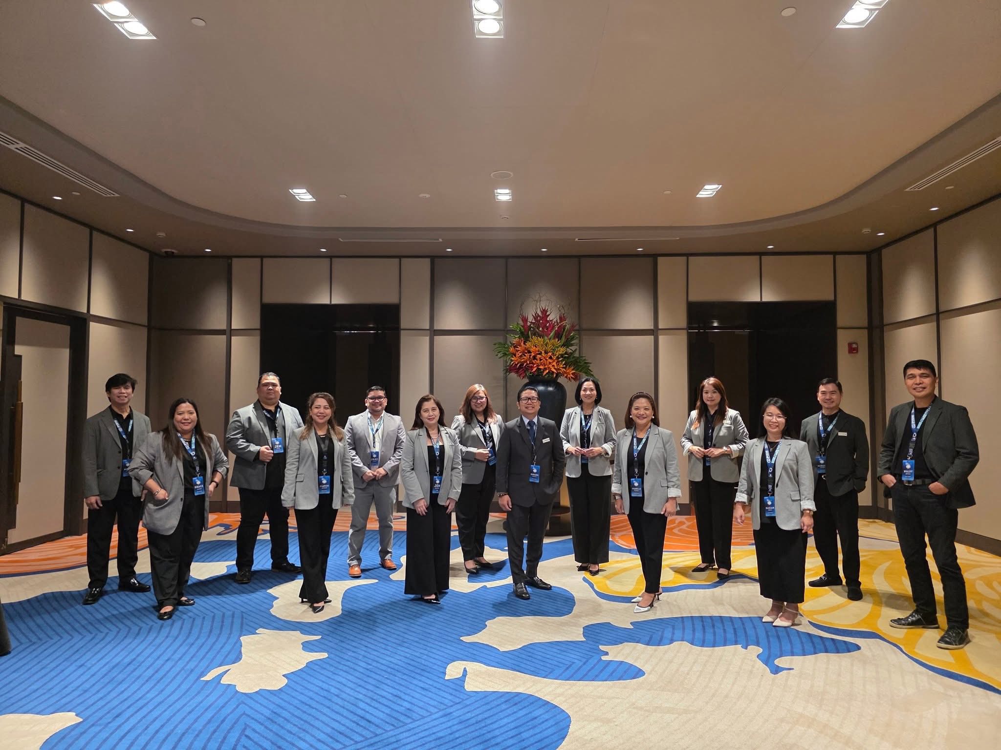 Group of professionally dressed individuals standing indoors on a colorful patterned carpet with a large floral arrangement behind them.