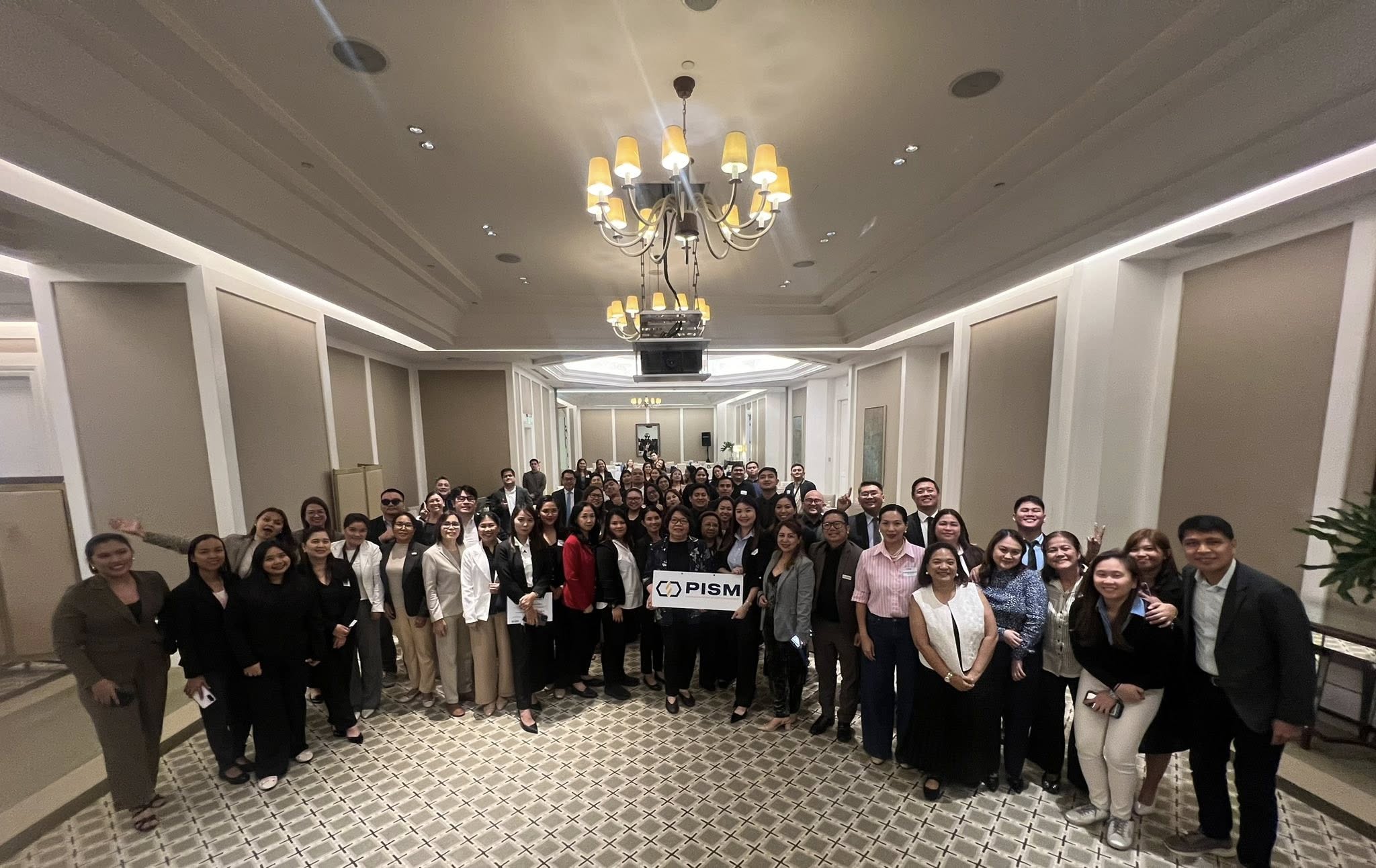 Large group of professionally dressed people posing indoors in a carpeted room holding a PISM sign.