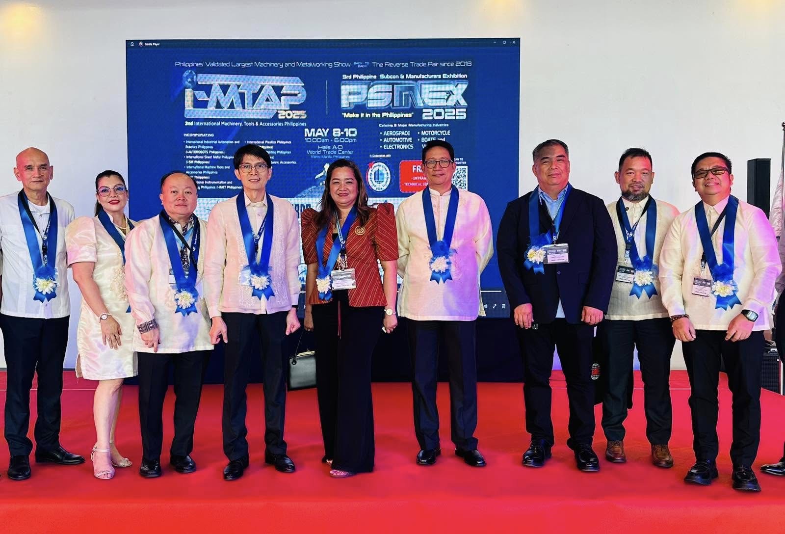 Group of nine people in formal Filipino attire standing on a red carpet with blue sashes and floral decorations around their necks, posing in front of a screen displaying logos for IMTAP 2025 and PESMEX 2025 events.