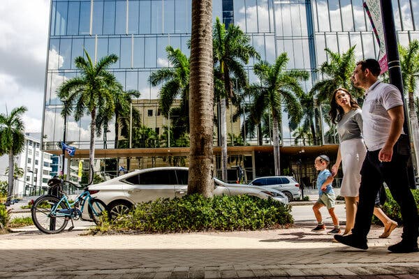 People walking along a sidewalk in front of a large building and palms trees.
