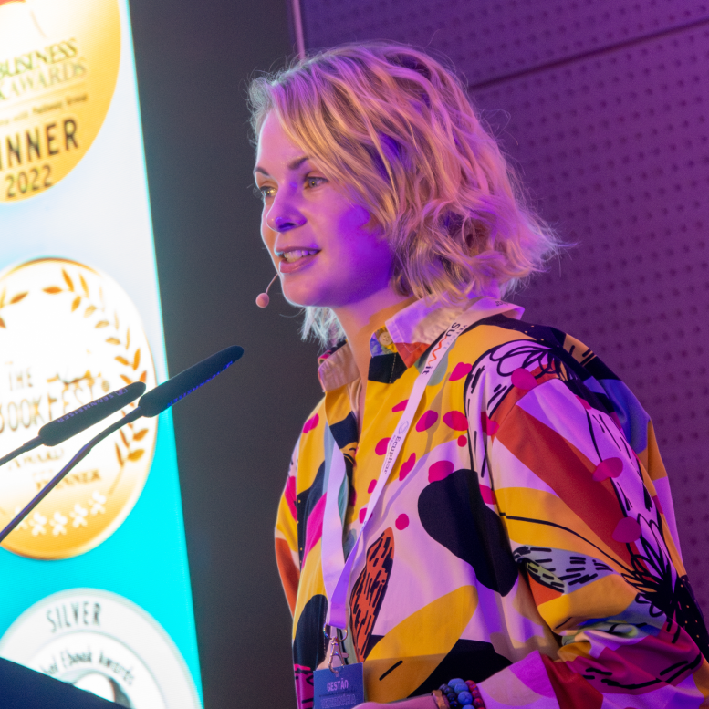 Jenna Tifanny wearing a colorful patterned shirt and a headset microphone, speaking at a podium with award logos visible in the background.