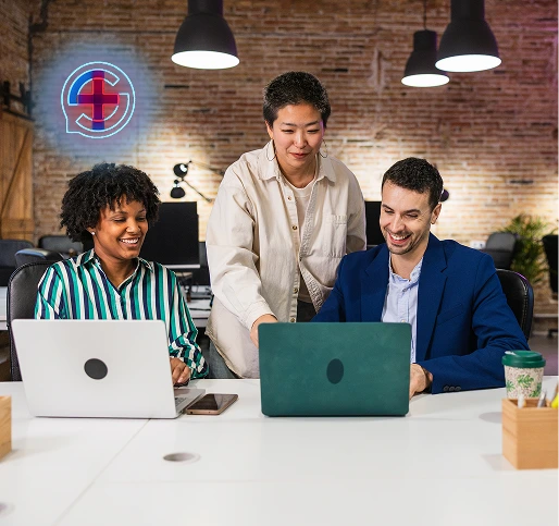 Three colleagues in a modern office with exposed brick wall and hanging lights, working together on laptops and smiling.