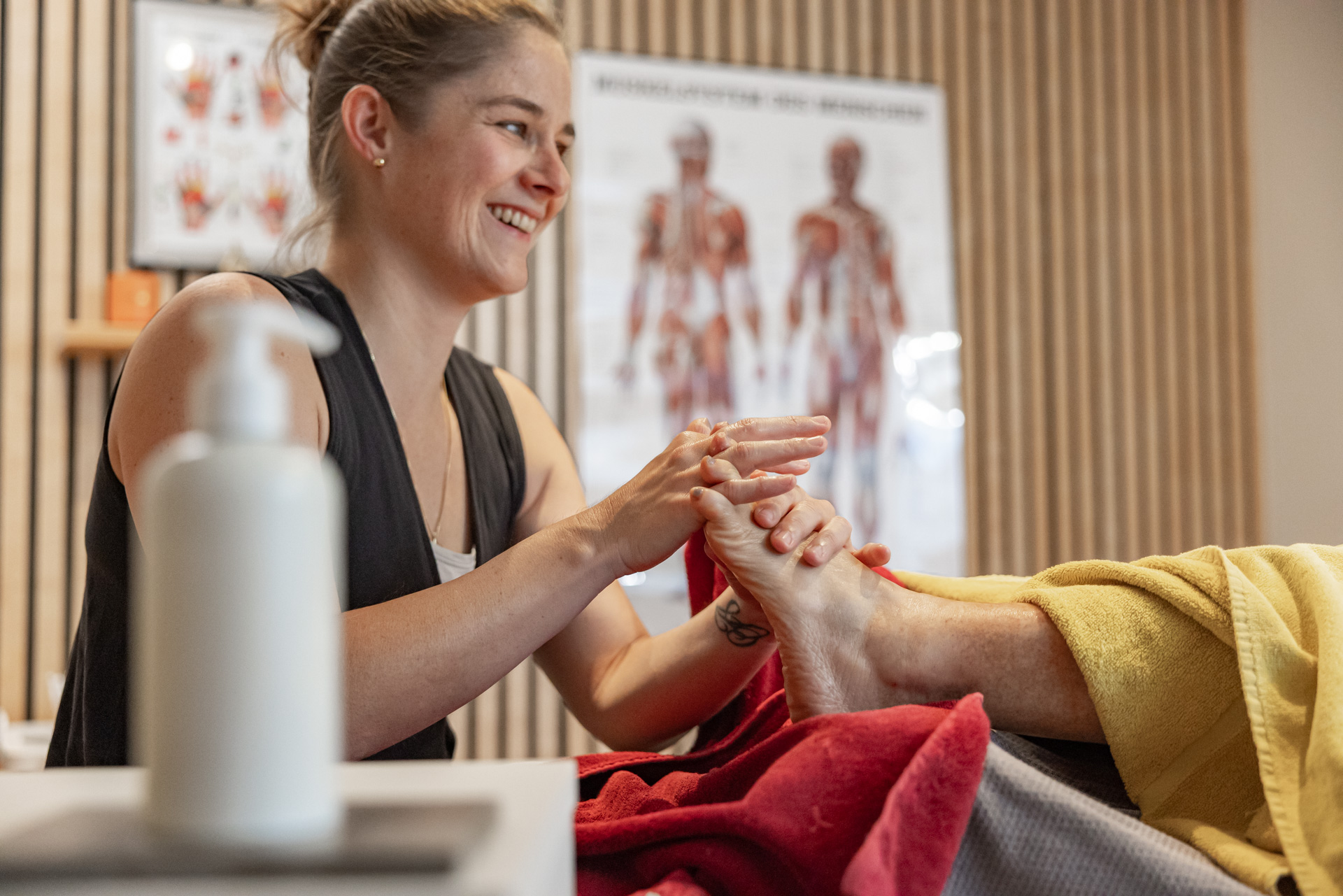 Die lächelnde Masseurin Angela Hoch von Hoch-Gefühl führt eine Fußmassage durch. Stimmungsvolles Bild einer entspannenden Behandlung in der Praxis in Diepoldsau mit Anatomie-Postern im Hintergrund.