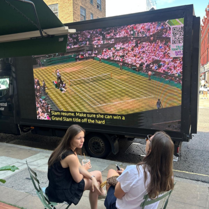 Two women sitting on sidewalk chairs watching a tennis match on a large outdoor screen mounted on a truck.