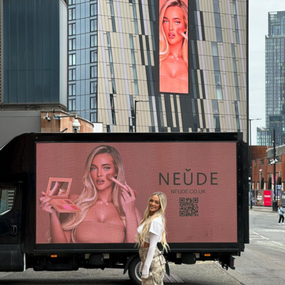Blonde woman standing in front of a truck with a pink advertisement for NEÜDE cosmetics featuring a model applying makeup.