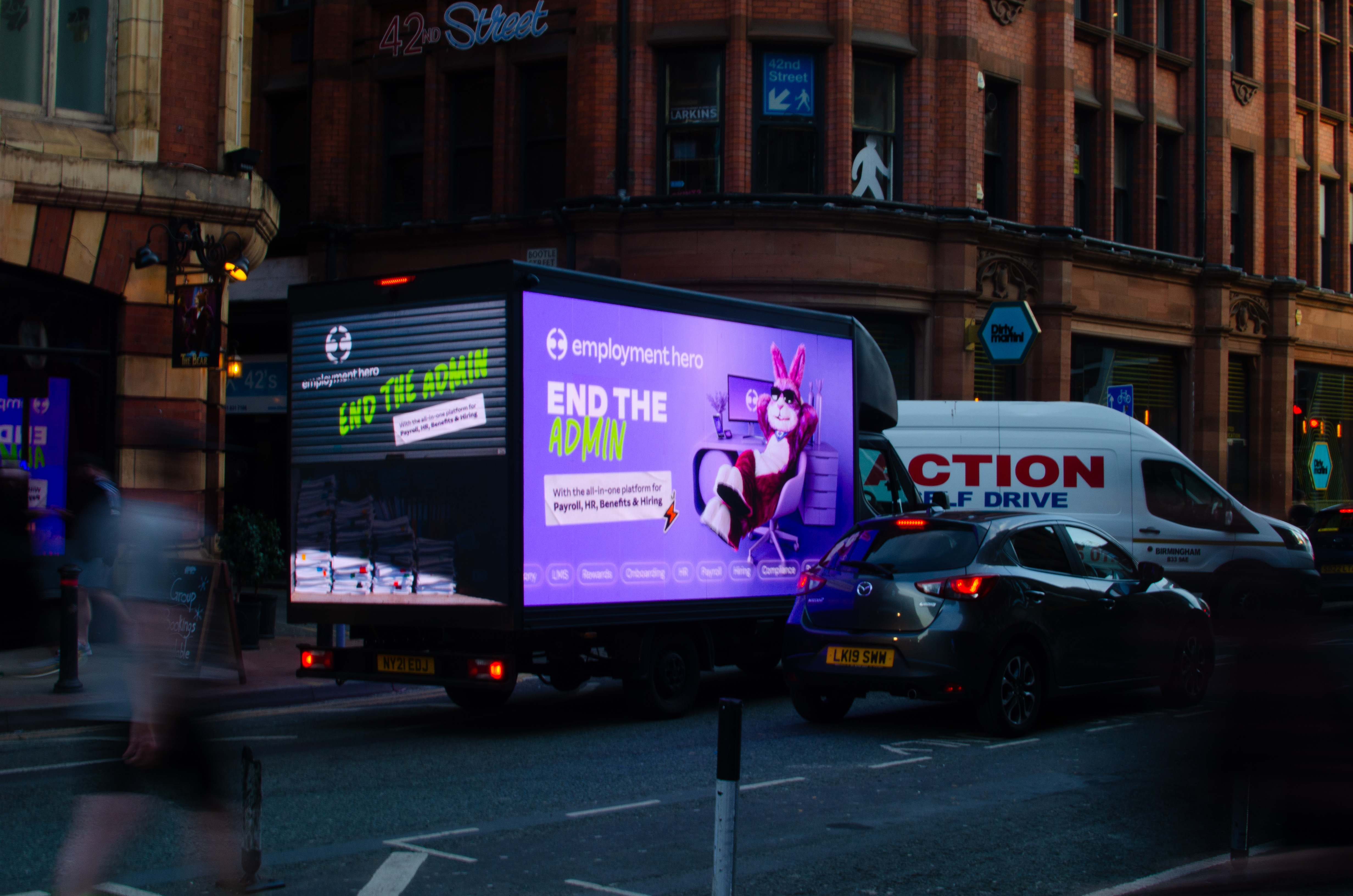 A street scene at dusk with a vehicle displaying a bright advertisement for Employment Hero saying 'End the Admin' featuring a person in a rabbit costume sitting in an office chair.