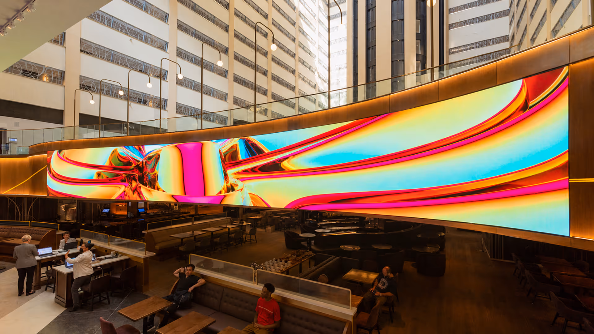 Modern indoor space with a large, colorful, curved digital screen displaying an abstract design above a lounge area with seated people.