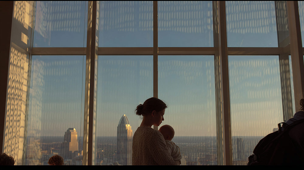 Silhouetted woman holding a baby in front of large windows with city skyline and patterned shadows at sunset.