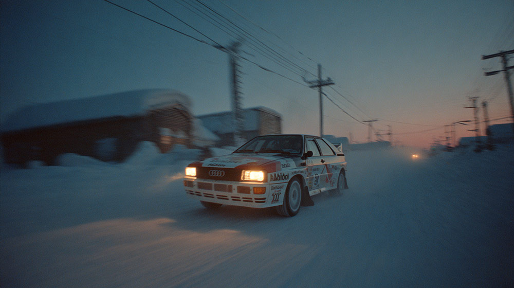 Audi Quattro rally car driving on a snowy road at dusk with headlights on and snow-covered buildings in the background.