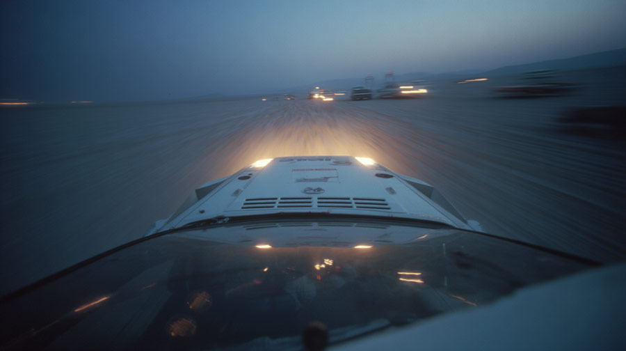 View from inside a vehicle driving fast on a sandy terrain at dusk with headlights illuminating the path and blurred lights ahead.