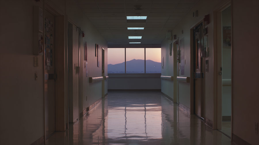 Dimly lit hospital corridor with polished floor reflecting ceiling lights and a sunset view of mountains through a window at the end.