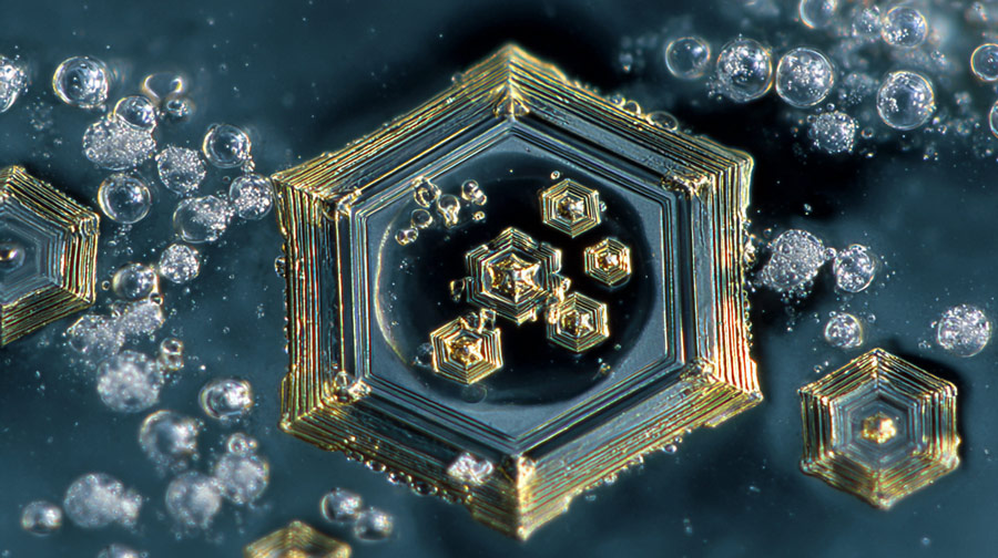 Close-up of intricate hexagonal crystalline structures with surrounding small bubbles on a dark surface.