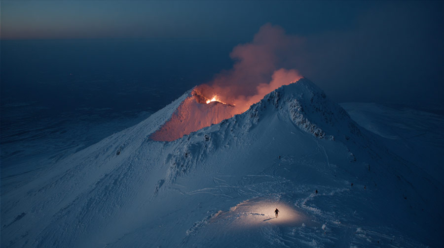 Snow-covered erupting volcano glowing with lava and smoke at night, with a lone person illuminated by a light standing nearby.