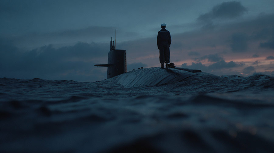 Person in naval uniform standing on a submarine hull at sea during dusk with dark clouds in the sky.