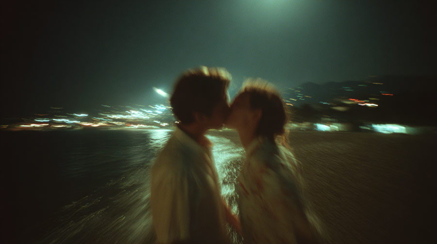 Blurry image of a couple kissing at night on a beach with city lights in the background.