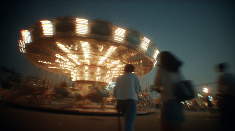Blurry nighttime scene of a brightly lit spinning swing ride at an amusement park with indistinct figures in the foreground.