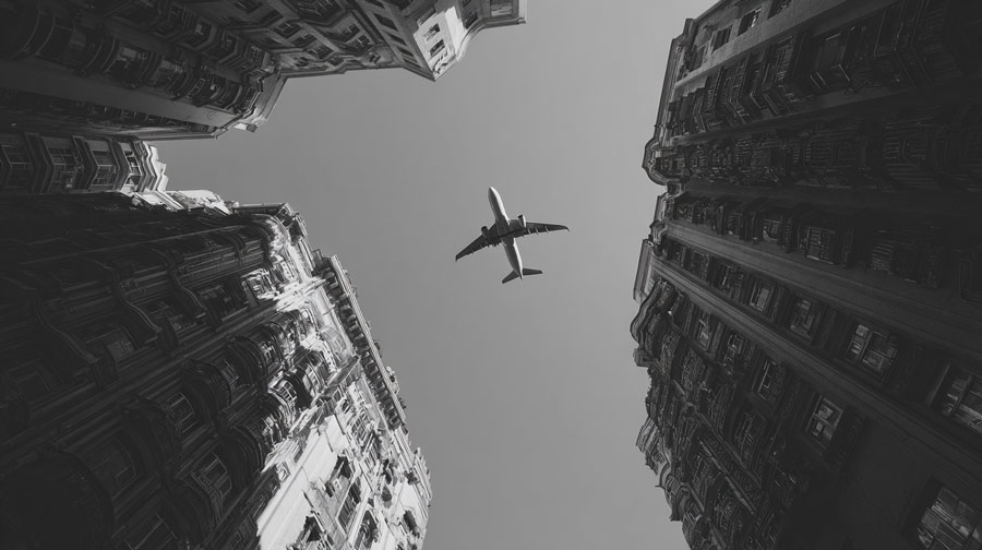 Airplane flying overhead framed by tall urban buildings from a ground-up perspective in black and white.