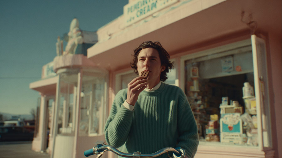 Young man in a teal sweater eating a chocolate ice cream bar while holding bicycle handlebars outside a pink ice cream shop.
