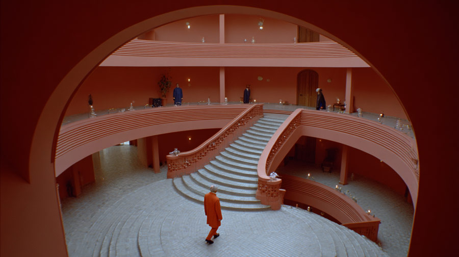 Interior of a spacious circular building with terracotta walls, featuring a wide central staircase and multiple levels with railings, and five people dressed in colorful clothes positioned around the space.