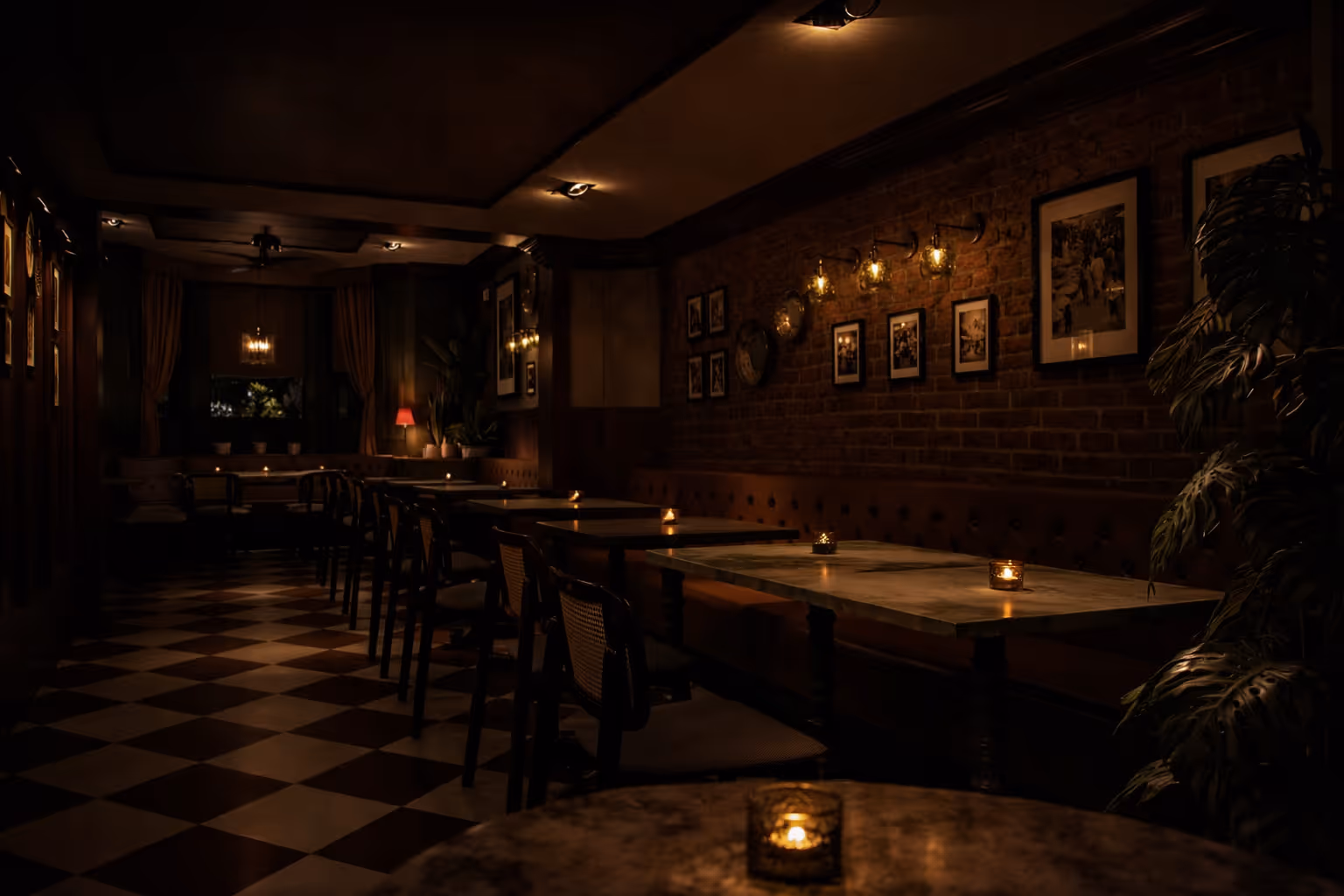 Dimly lit restaurant interior with checkered floor, rows of tables, chairs, candles, and framed pictures on a brick wall.