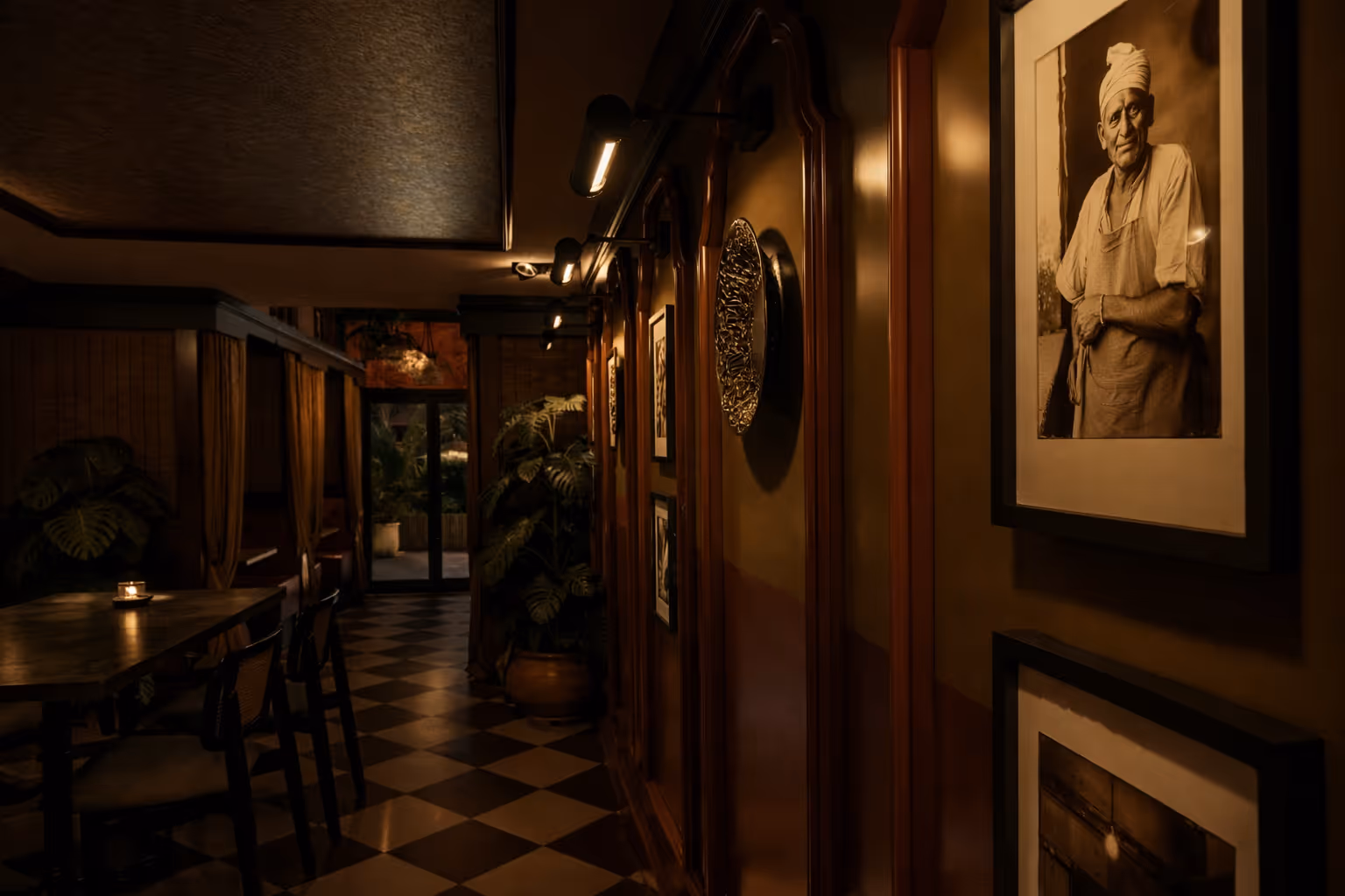 Dimly lit hallway with framed black-and-white portrait of a man in an apron and head wrap on the right wall, wooden paneling, checkered floor, and a table with chairs and a lit candle on the left.