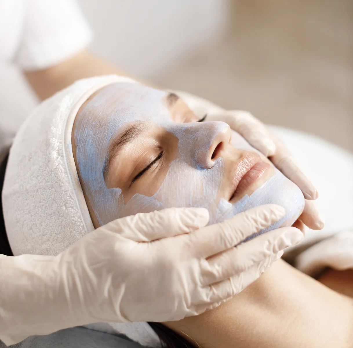 Woman with a white facial mask receiving a facial treatment while wearing a white headband and closed eyes.