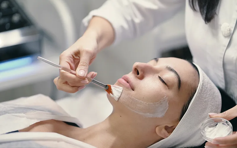 Woman receiving a facial treatment with a brush applying cream on her cheek while lying down with eyes closed.