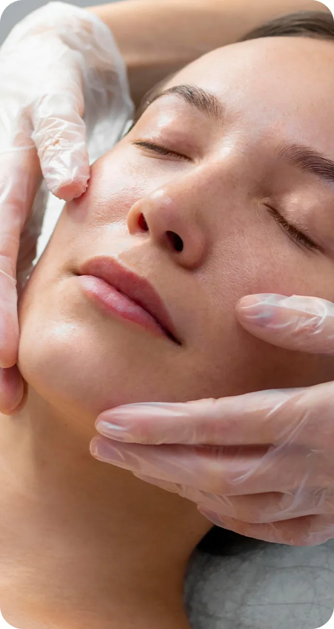 Close-up of a woman with eyes closed receiving a facial treatment from hands wearing medical gloves.