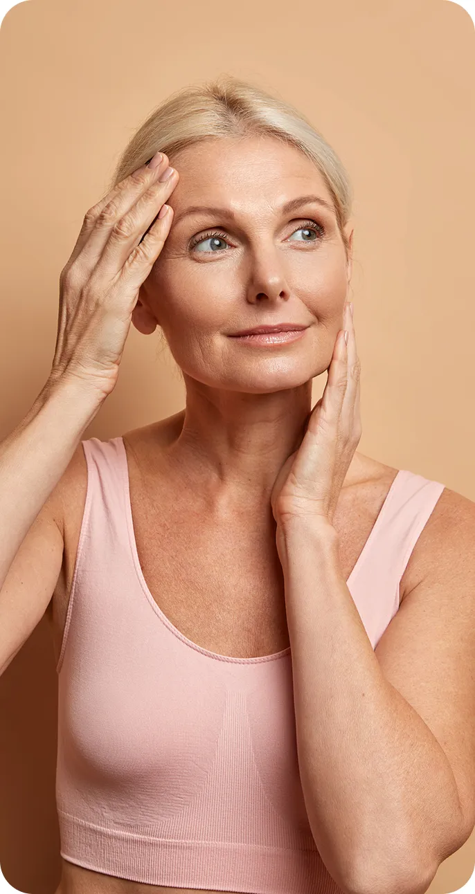 Mature woman in a pink tank top gently touching her face and forehead with a thoughtful expression.