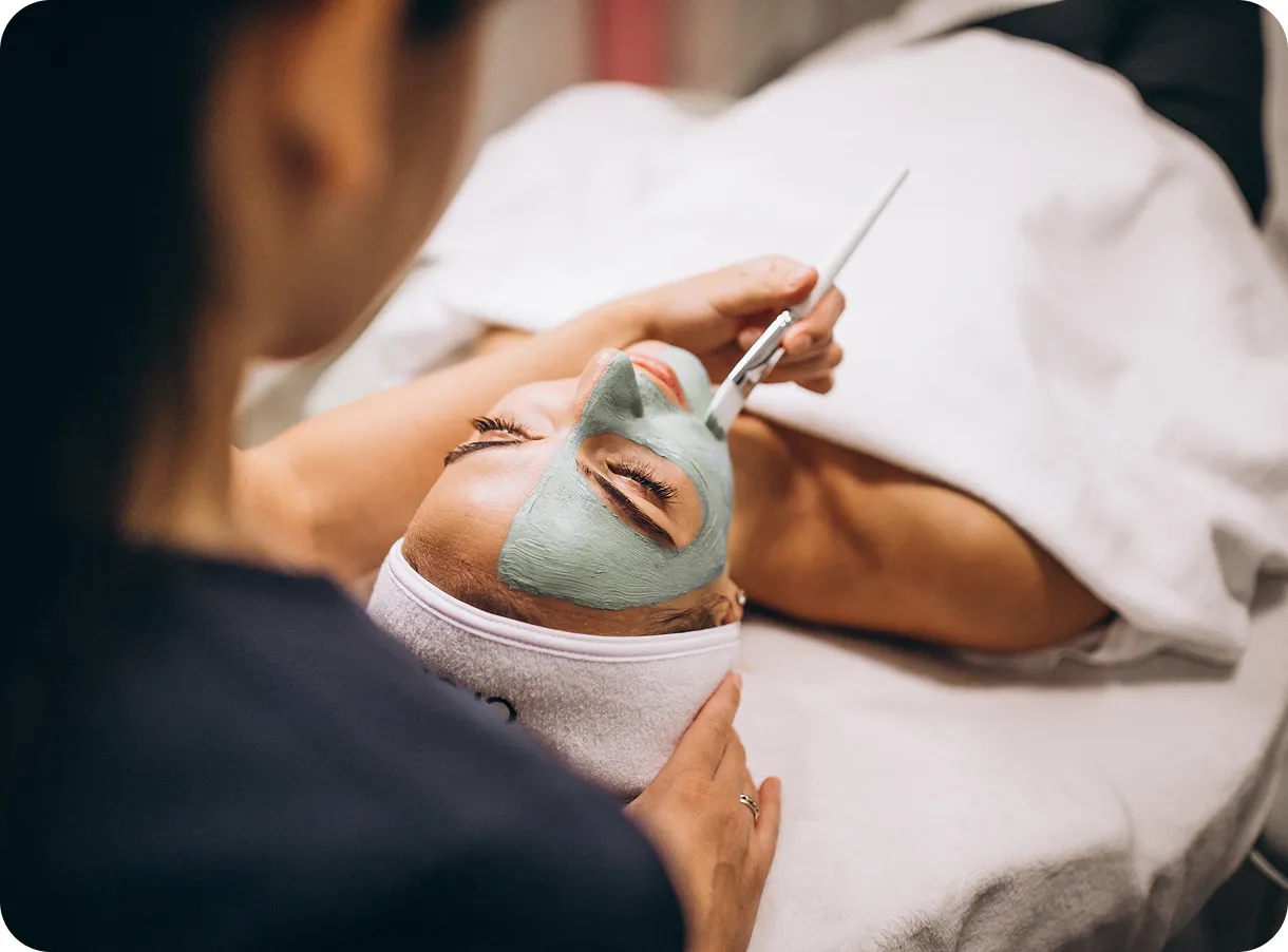 Woman with eyes closed receiving a green facial mask application while lying on a spa bed.