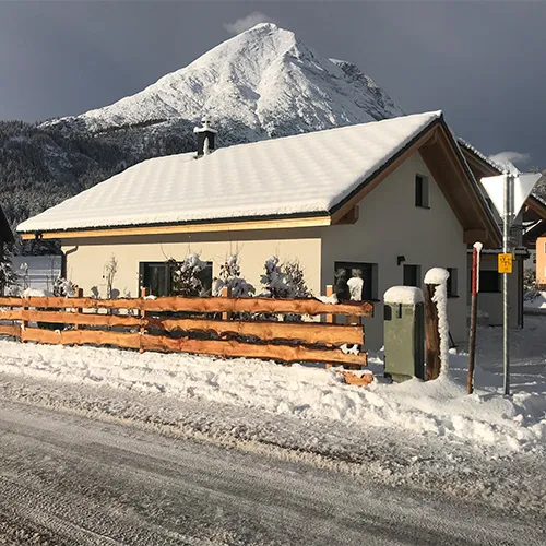 Verschneites Chalet mit Holzzaun und Berglandschaft im Hintergrund unter bewölktem Himmel.