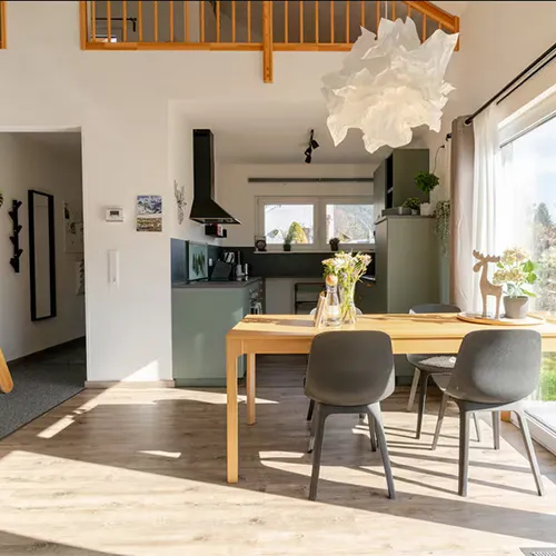 Heller, moderner Essbereich mit Holztisch, grauen Stühlen und großem Fenster mit Sonneneinfall, angrenzend an eine kompakte Küche.